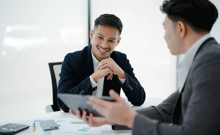 100+ Sales Manager Interview Questions With Answers Evinex Corporation | Staffing Agency a smiling man with his hands together looking at a tablet that a man is holding