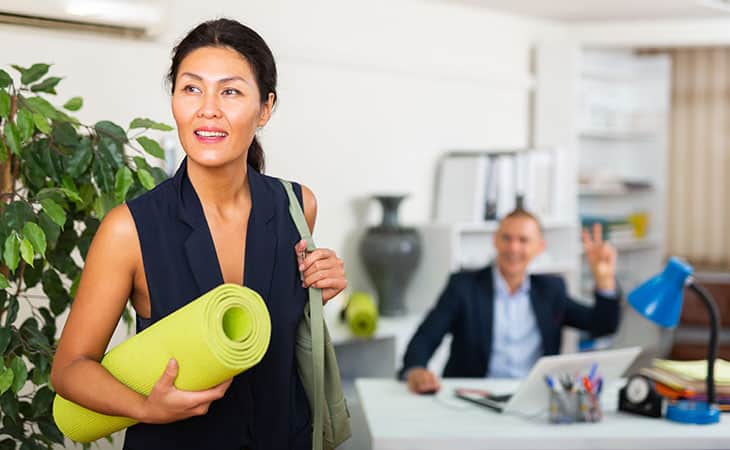 Hr Issues And Challenges: Solutions For The Modern Workplace Evinex Corporation | Staffing Agency a woman holding a yoga mat while walking past a colleague who is waving in the office