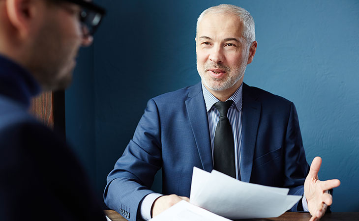 How To Impress In An Interview: Tips To Stand Out Evinex Corporation | Staffing Agency a businessman holding papers and an open hand talking to someone