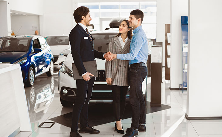 a businessman shaking hands with a man next to a woman in a car dealership