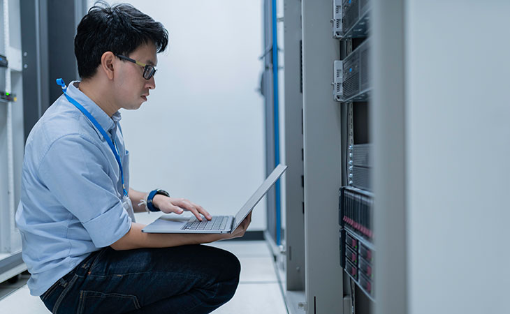 a crouching man using a laptop in a server room