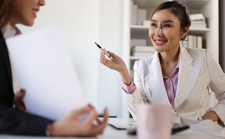 How To Impress In An Interview: Tips To Stand Out Evinex Corporation | Staffing Agency a smiling woman holding a pen while talking to a woman