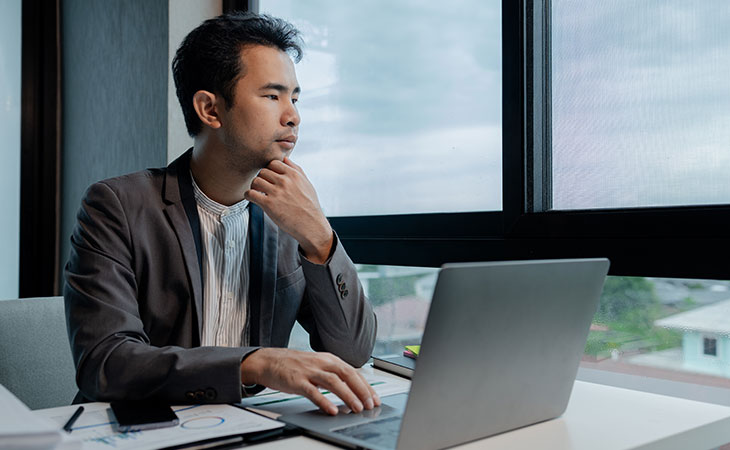 a thoughtful man looking outside the window with his hand on his chin and using a laptop