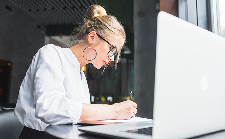 How To Impress In An Interview: Tips To Stand Out Evinex Corporation | Staffing Agency a woman writing with a pen on paper next to a laptop