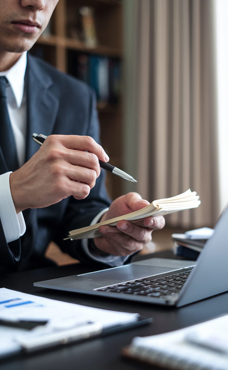 detail of a man holding a pen while cheking his notes in front of a laptop